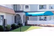 Front entrance of a light-gray rehabilitation and health care center with a blue awning, white picket fence, and surrounding shrubs.