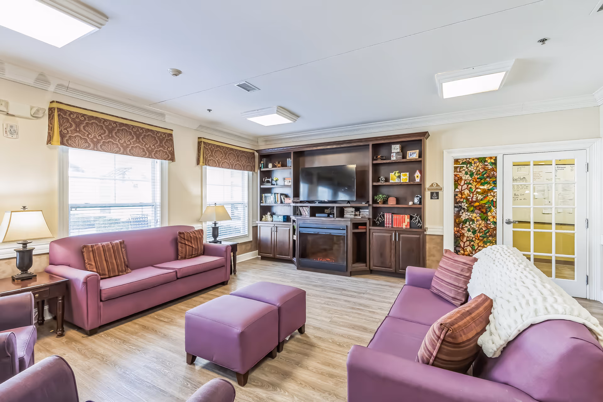 A bright and cozy living room with two purple sofas, a matching ottoman, and a dark wooden entertainment center with a flat-screen TV and electric fireplace. The room has large windows with patterned valances, two table lamps, and a door with a colorful stained glass design.