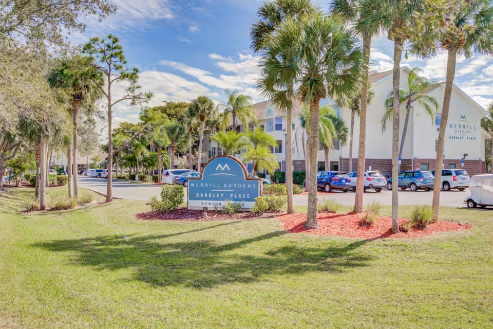 Exterior view of Merrill Gardens at Barkley Place senior living facility with a landscaped lawn, palm trees, a parking lot with cars, and a clear blue sky. The facility's sign is prominently displayed in the foreground.