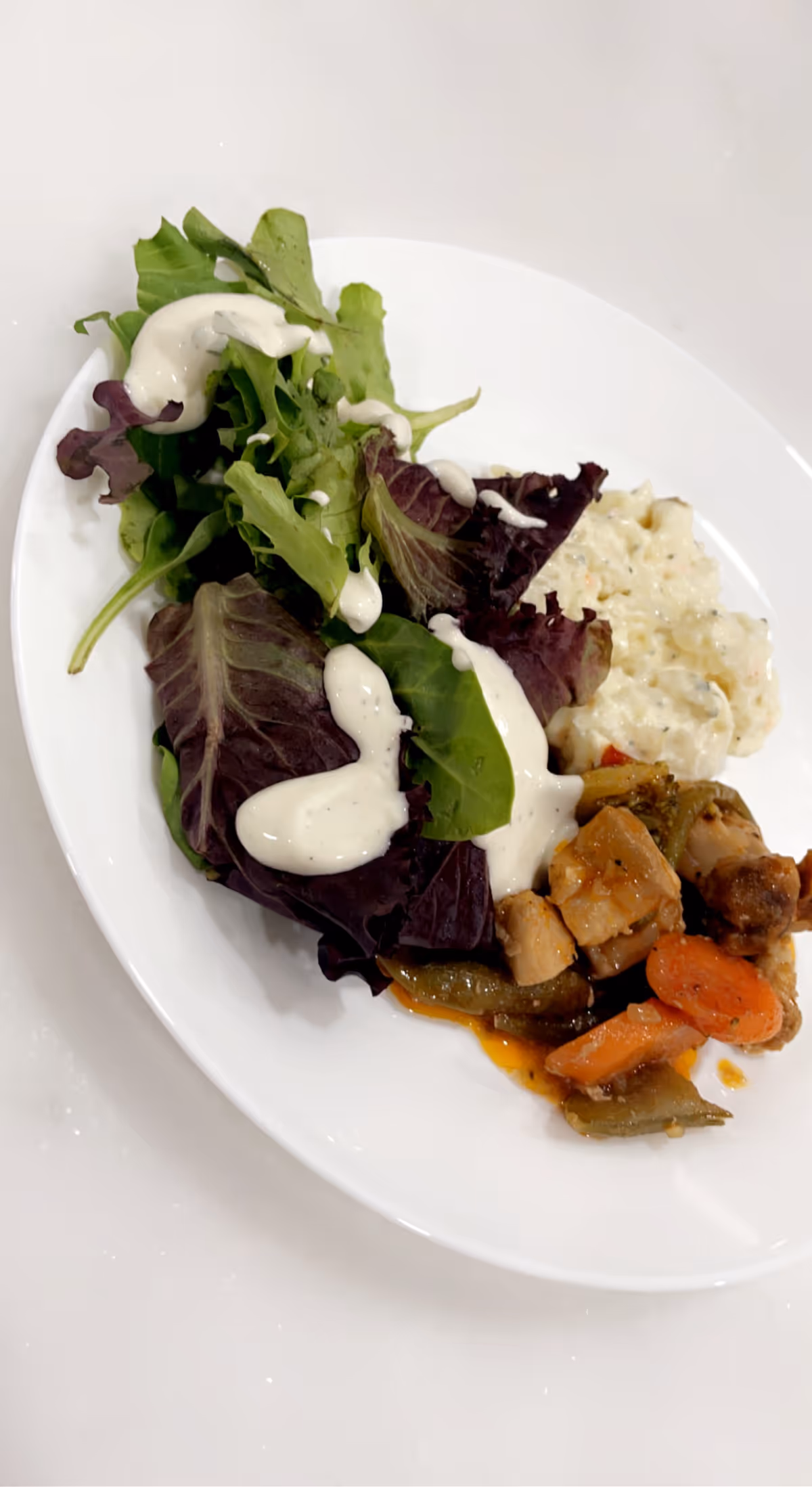 A white plate with a serving of mixed green salad topped with creamy dressing, a portion of creamy potato salad, and cooked vegetables including carrots and green beans with pieces of chicken.