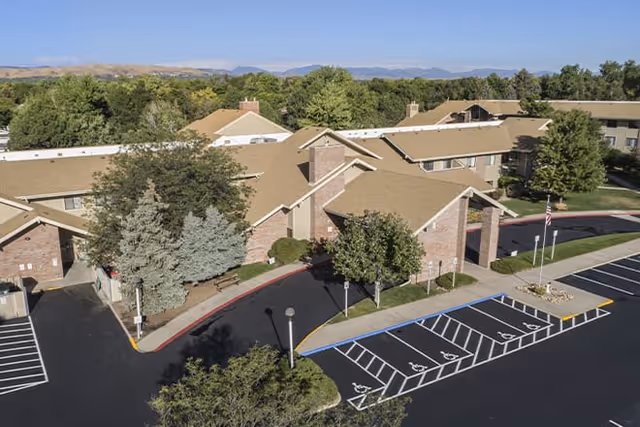 Aerial view of Brookdale Meridian Lakewood senior living facility showing multiple connected buildings with beige roofs surrounded by trees and greenery. There is a parking lot with several handicap parking spaces in the foreground and mountains visible in the background under a clear blue sky.