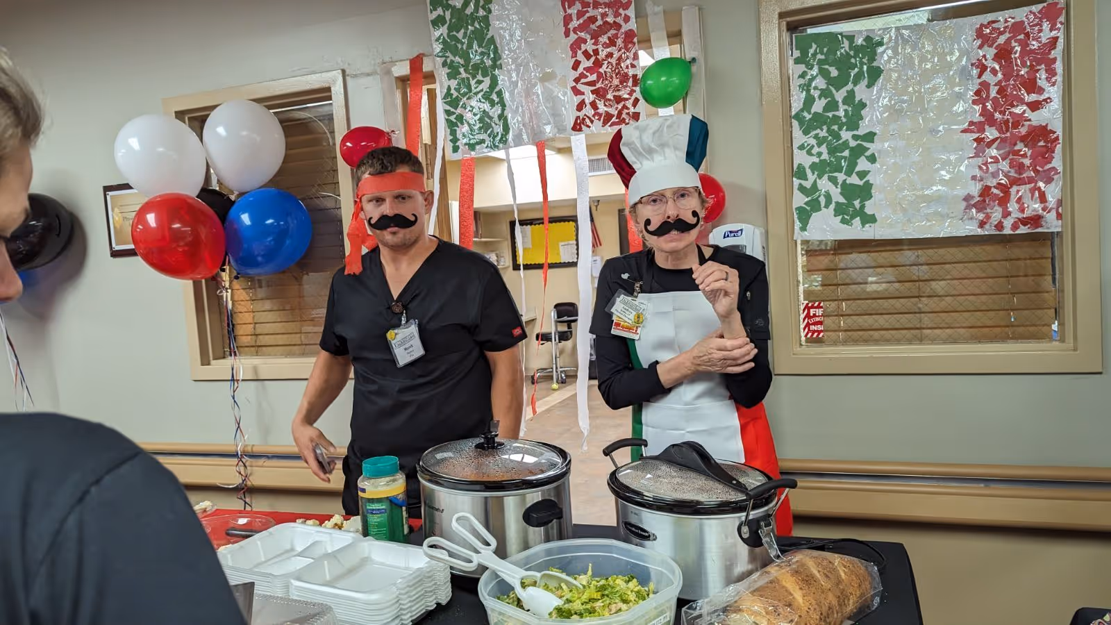 Two staff members dressed in black scrubs and Italian-themed costumes, including chef hats and fake mustaches, standing behind a table with food containers and a salad bowl. The room is decorated with red, white, and green balloons and streamers, and Italian flag decorations are visible on the windows.