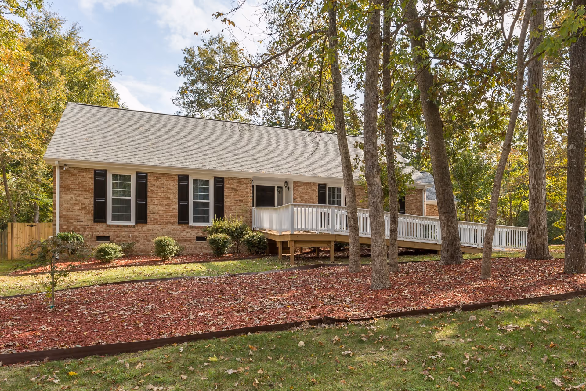 Exterior view of a single-story brick building with a gray shingled roof and black window shutters. The building has a white wooden ramp leading to the front door. The surrounding area features trees, green grass, and a landscaped area with red mulch and small bushes.