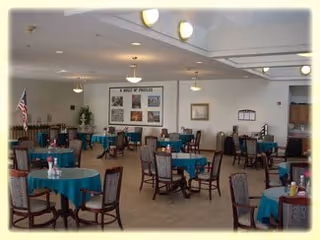 A spacious dining room with multiple round tables covered with teal tablecloths, each surrounded by wooden chairs with cushioned seats. The room has beige walls with framed pictures and an American flag in the corner. The ceiling has recessed lighting and hanging light fixtures.