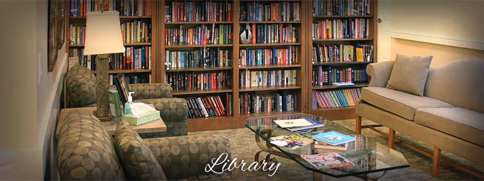 Cozy library room with bookshelves filled with books along the back wall, two patterned armchairs, a beige sofa with cushions, a glass coffee table with magazines and books, and a floor lamp providing warm lighting.