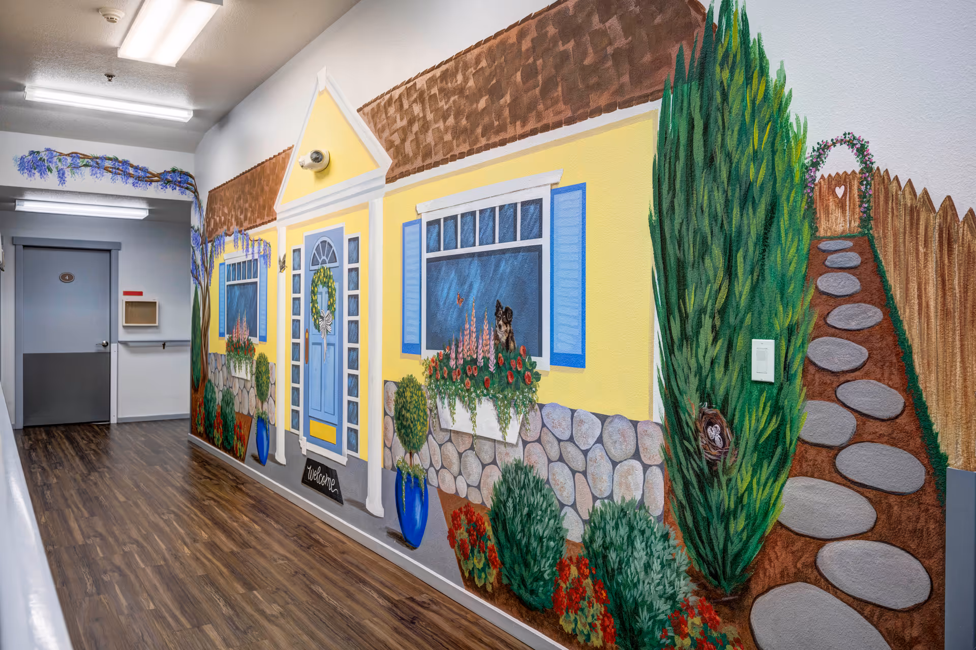 A hallway with wood-patterned flooring and white walls, featuring a large mural on one side depicting a yellow house with blue shutters, a blue door with a wreath, flower boxes, bushes, and a stone pathway leading to a wooden gate with a heart-shaped cutout. The ceiling has fluorescent lights, and there is a gray door at the end of the hallway.