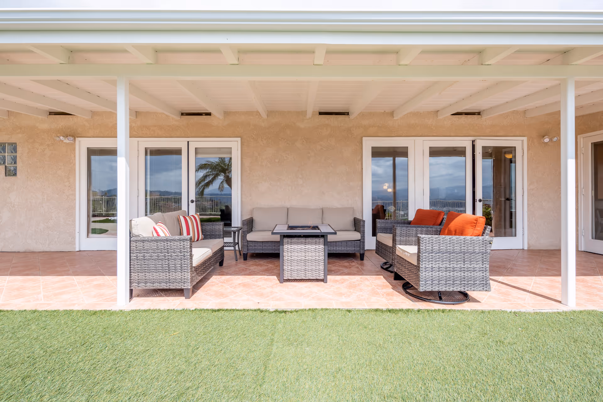 Outdoor covered patio area with wicker furniture including a sofa, two armchairs with orange cushions, and a coffee table on a tiled floor, overlooking a green lawn with a view of palm trees and mountains in the distance.