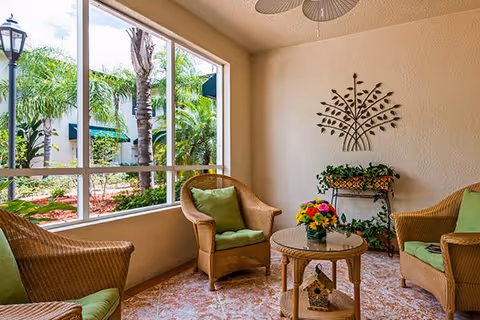 A cozy indoor seating area with three wicker chairs featuring green cushions, a round glass-top wicker table with a colorful flower arrangement, and a decorative metal wall art piece above a plant stand with greenery. Large windows provide a view of palm trees and greenery outside.