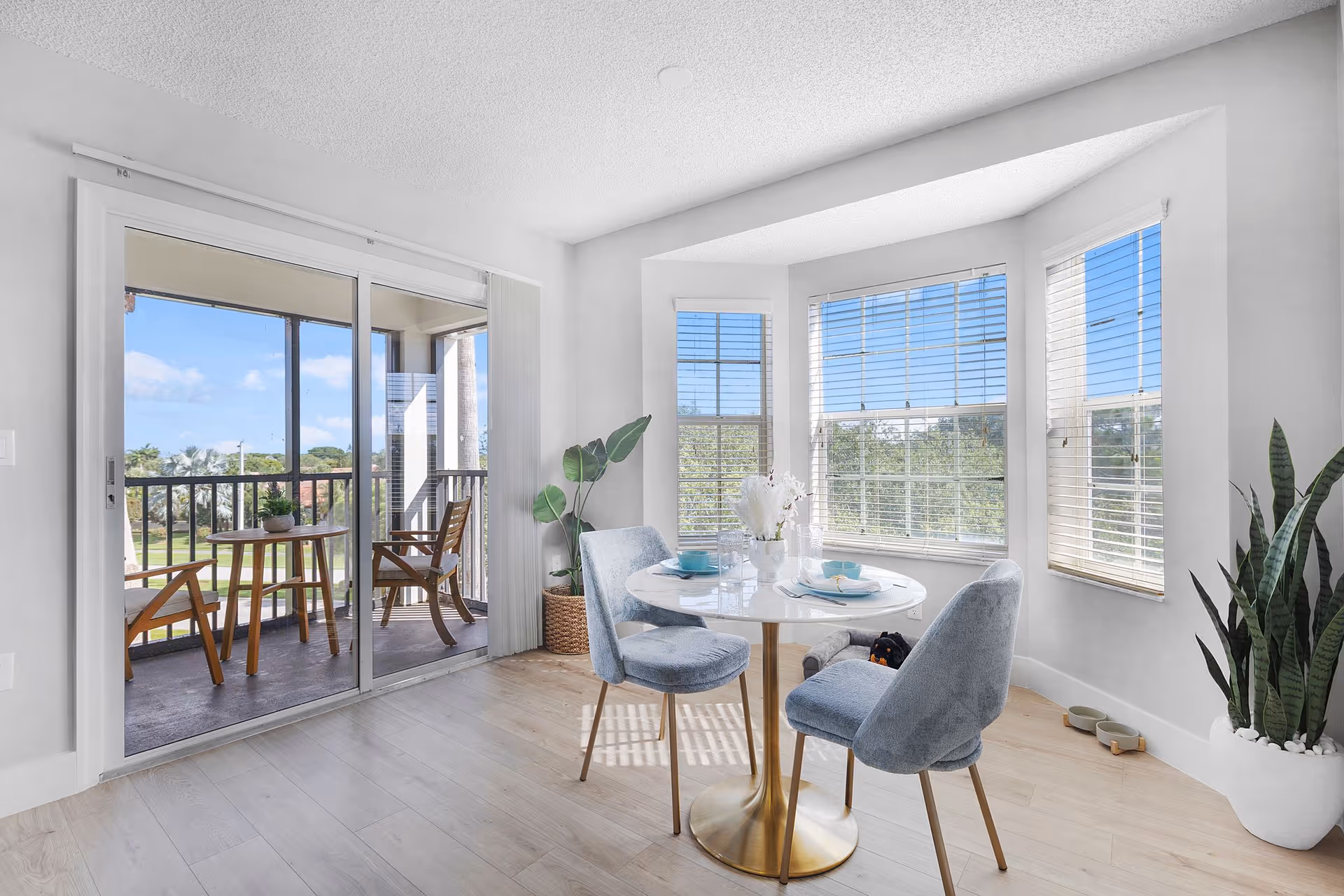 Bright dining nook with a round table and three upholstered chairs by bay windows and sliding doors leading to a balcony with outdoor seating.