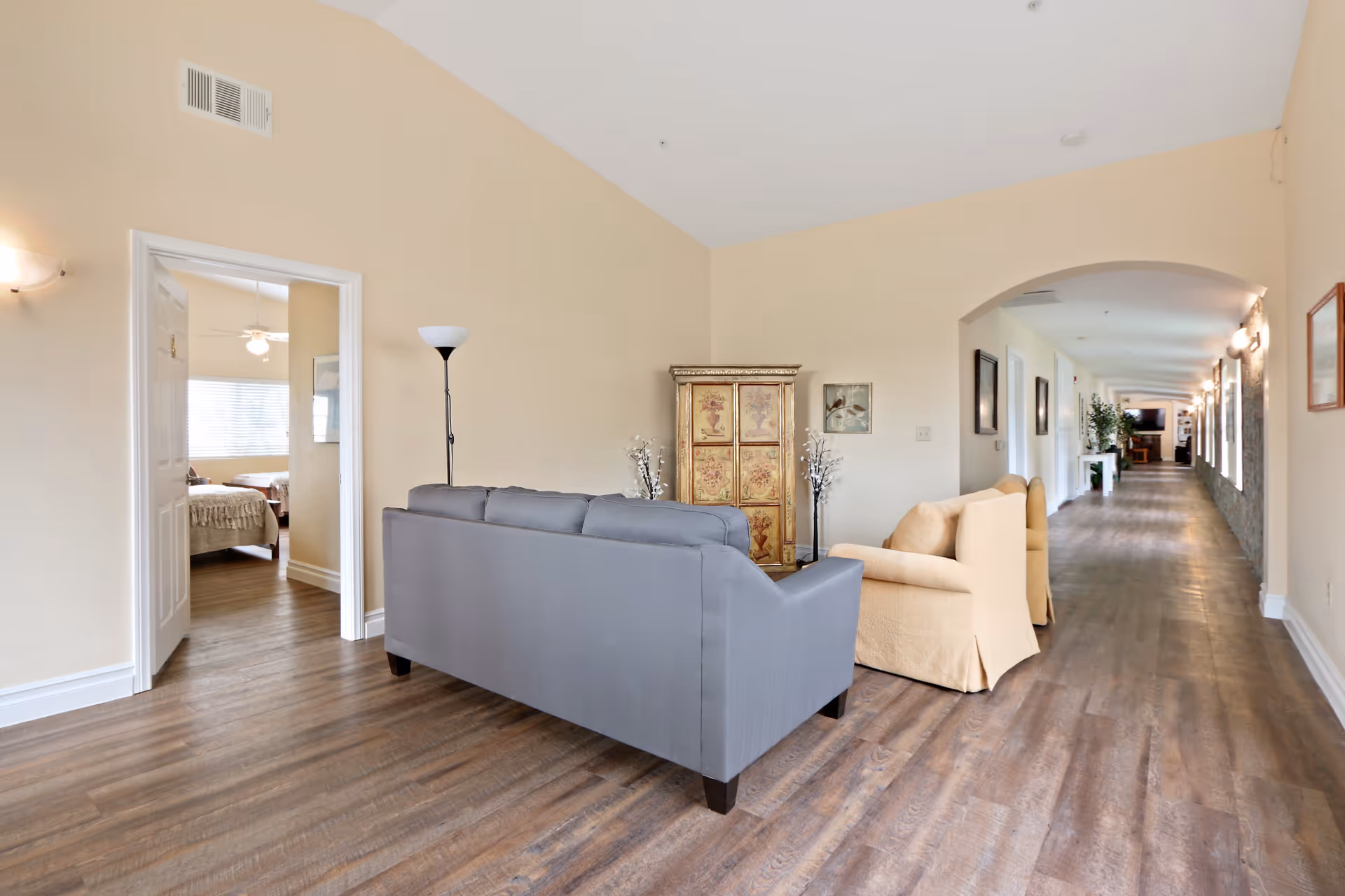 A spacious interior hallway in a senior living facility with wooden flooring and beige walls. The hallway features a seating area with a gray sofa and two beige armchairs, a decorative cabinet, and a floor lamp. To the left, an open door reveals a bedroom with a bed and a ceiling fan. The hallway extends into the distance with framed pictures on the walls and additional seating and plants visible further down.