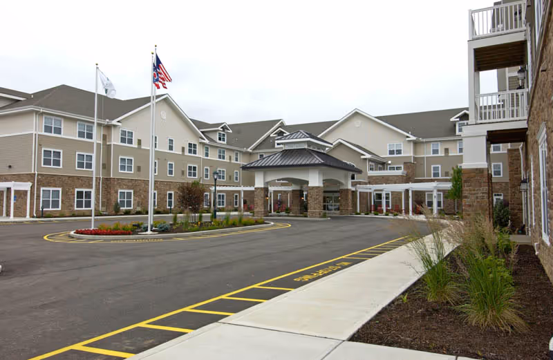 Exterior view of Pinebrook of Milford by StoryPoint Group, showing a large multi-story building with beige and stone facade, a covered entrance with pillars, three flagpoles with flags, and landscaped areas with plants and flowers along the driveway and sidewalk.
