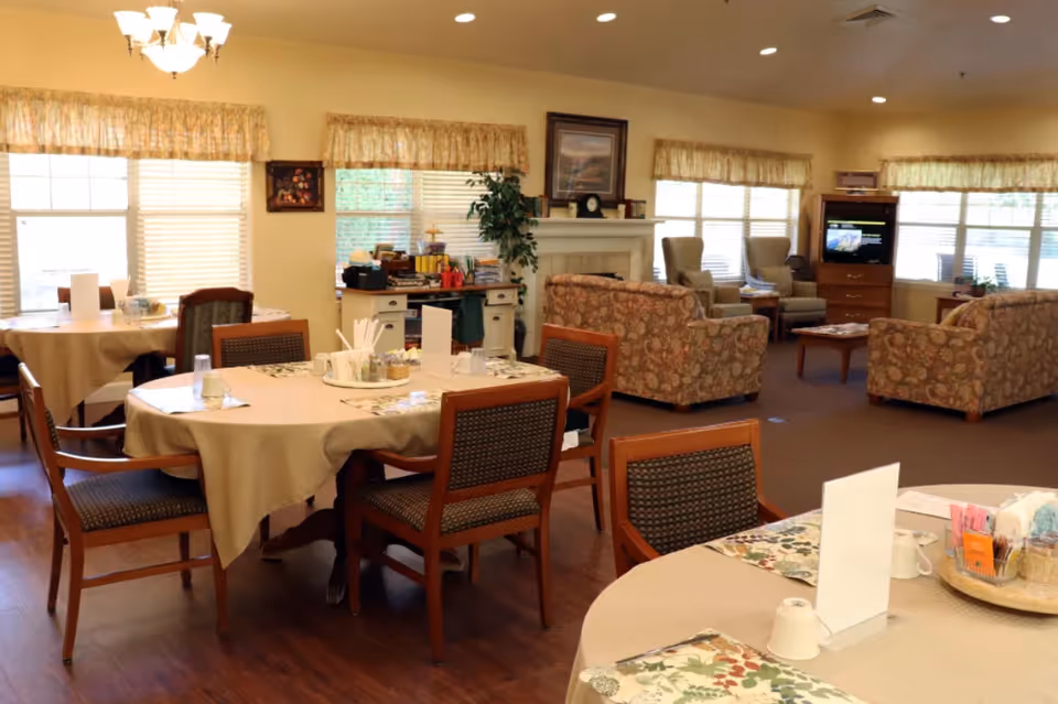A bright communal dining and sitting area with round tables set for meals, wooden chairs, upholstered sofas, and a television.