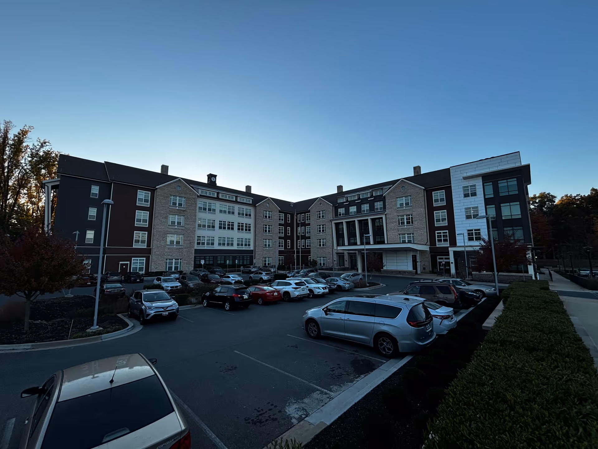 Front exterior of a multi-story senior living building with a parking lot and cars at dusk.