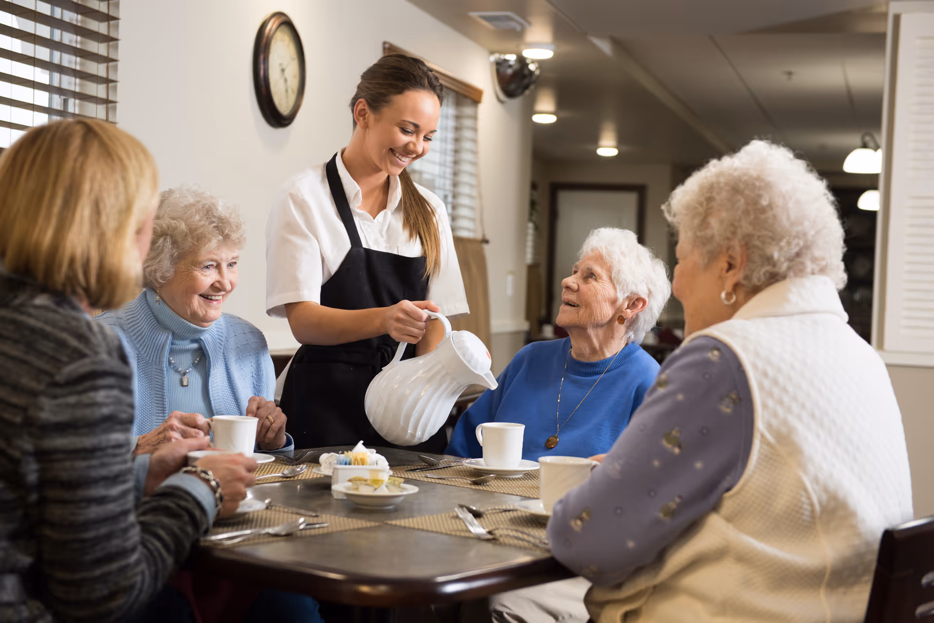 A caregiver pours coffee for a group of elderly women seated around a dining table in a communal dining room.