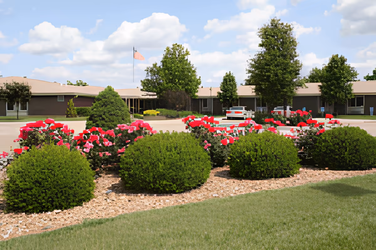 A landscaped outdoor area in front of a single-story building with a beige roof. The foreground features neatly trimmed green bushes and vibrant red flowers planted in a mulched bed. Several trees are visible around the building, and an American flag is flying on a flagpole near the center. The sky is partly cloudy with blue patches.