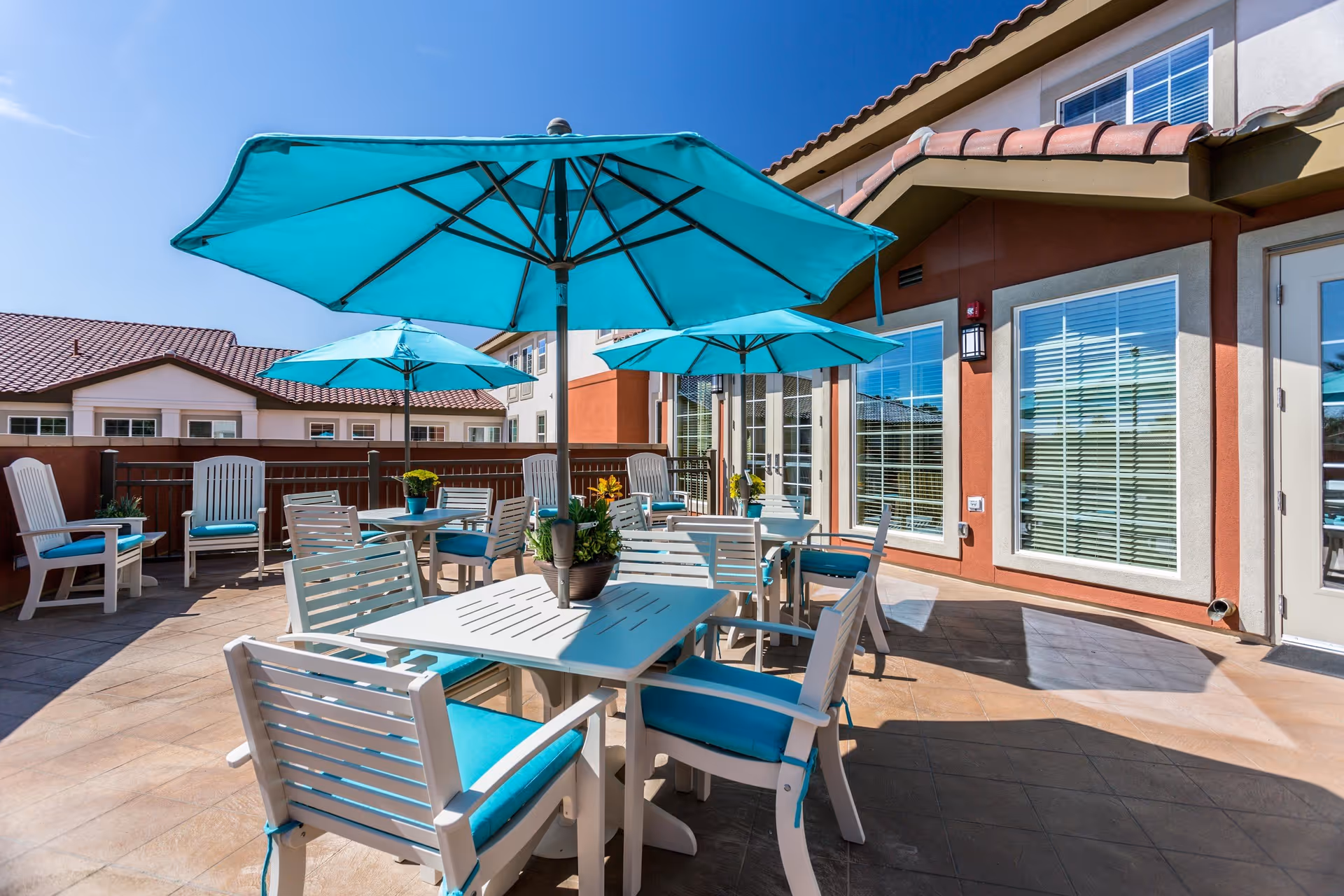 Sunny outdoor patio with white tables, turquoise umbrellas and chairs outside a residential building.