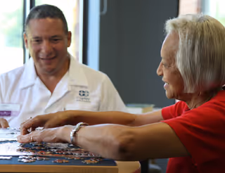 An elderly woman and a healthcare worker sitting at a table working on a jigsaw puzzle together in a well-lit room.