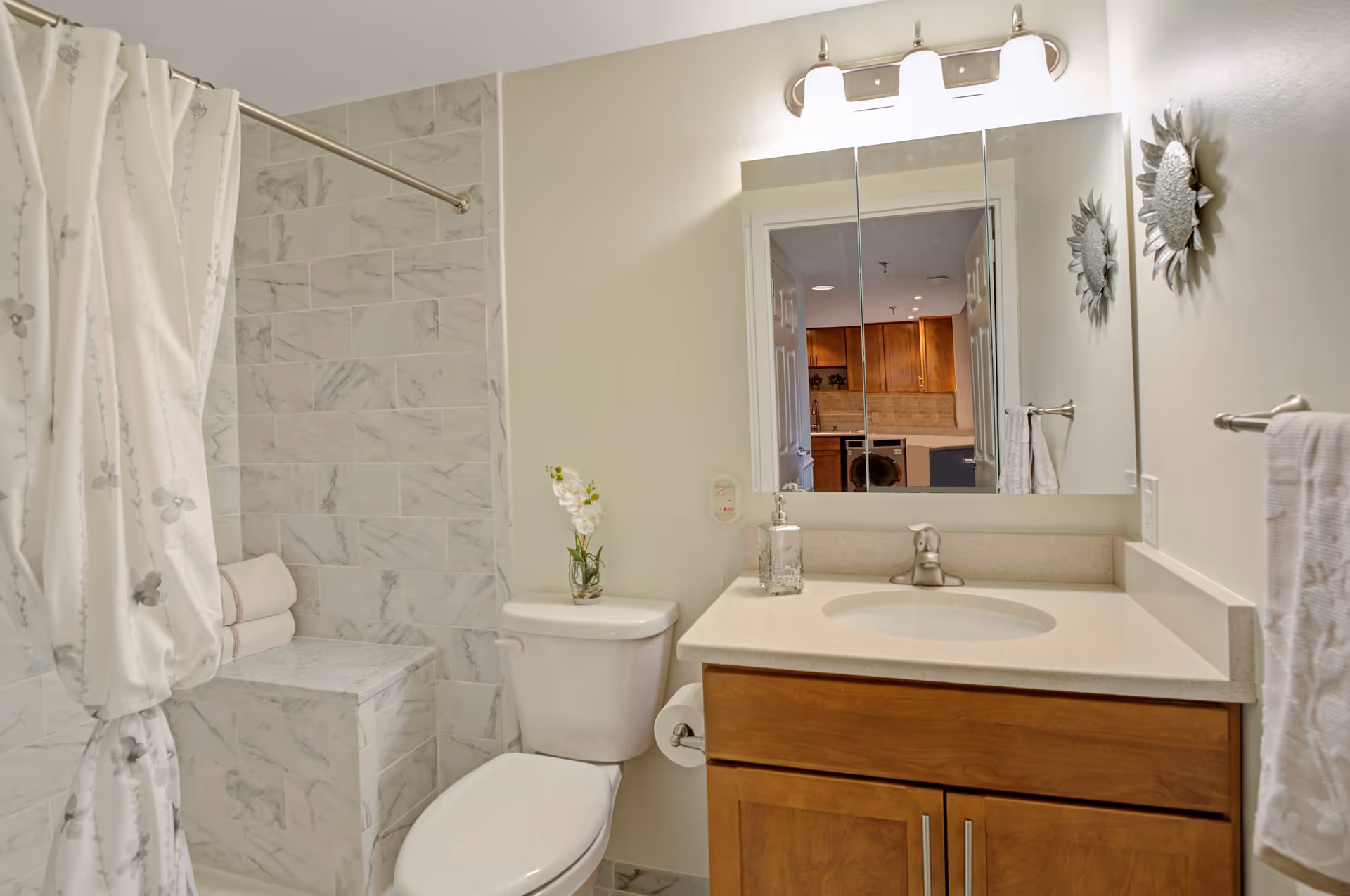 A clean and modern bathroom featuring a white toilet, a vanity with a sink and wooden cabinets, a large mirror with three panels, and a shower area with marble-like tiled walls and a white floral shower curtain. A small vase with white flowers is placed on the toilet tank, and a towel hangs on a rack to the right.