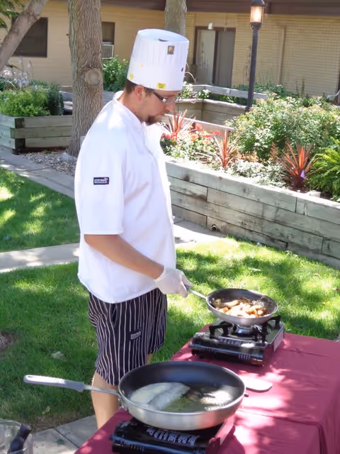 A chef in a white coat and tall hat cooks on portable burners at a table in a landscaped courtyard.