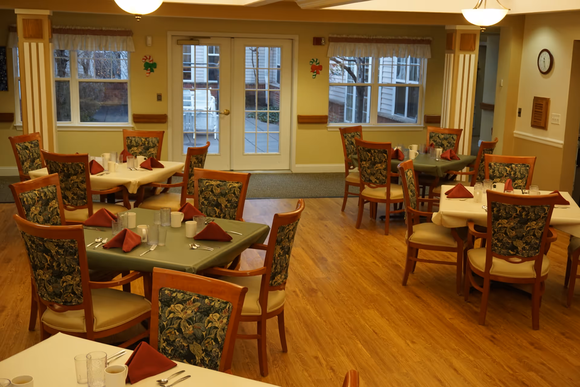 Dining room with multiple set tables and floral-upholstered chairs in a senior living facility.