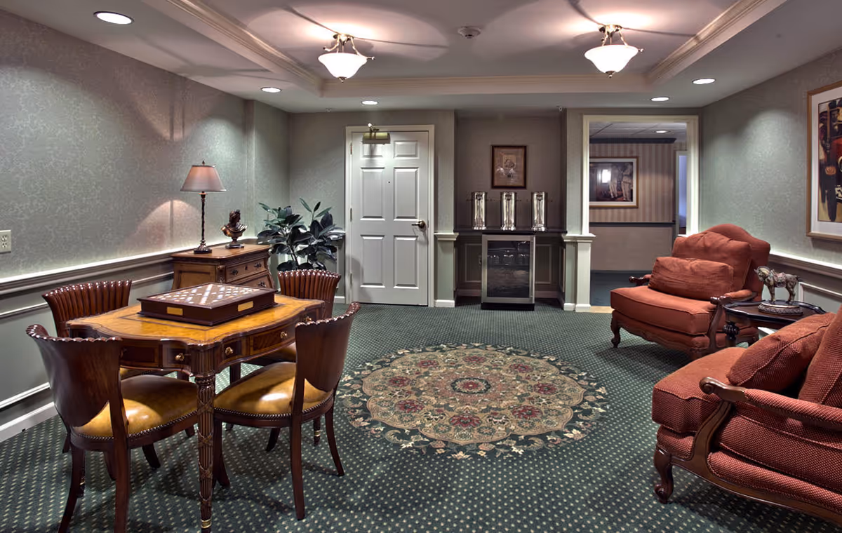 A cozy sitting area in a senior living facility featuring a round wooden table with four chairs, a chess/checkers board on the table, a wooden sideboard with a lamp and plant, two upholstered armchairs with cushions, a small side table with a decorative horse figurine, patterned carpet with a circular floral design, and framed artwork on the walls.