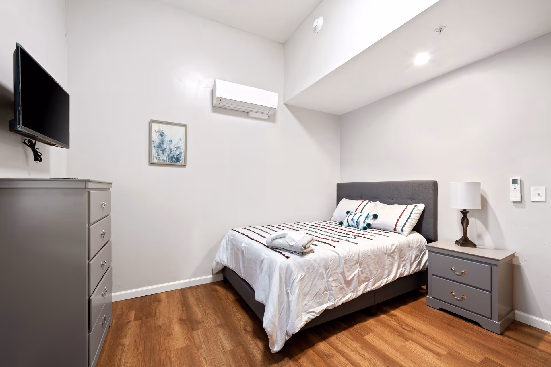 A neatly arranged bedroom with a bed featuring a white comforter with colorful stripes, a gray upholstered headboard, and pillows. There is a gray nightstand with a lamp on the right side of the bed and a gray chest of drawers on the left side of the room. A flat-screen TV is mounted on the left wall above the chest of drawers. The walls are painted white, and the floor has a wood finish. A small framed artwork and an air conditioning unit are mounted on the wall above the bed.