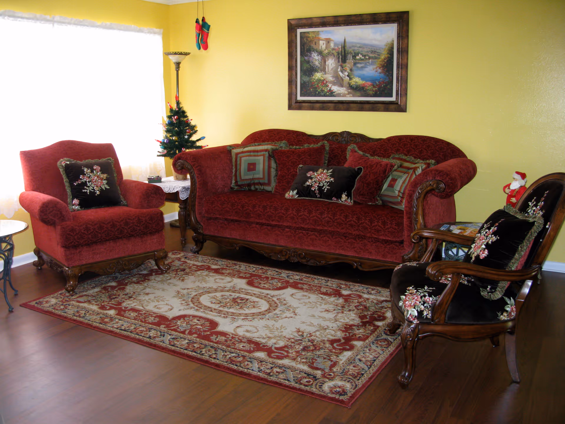 A cozy living room with a red upholstered sofa and armchair, both with decorative pillows. There is a wooden framed armchair with floral cushions, a patterned area rug on a wooden floor, a small Christmas tree on a side table, and a framed landscape painting on a yellow wall.