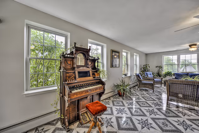 Bright sunroom-style living area featuring an ornate antique pump organ, patterned tile floor, wicker seating, plants, and multiple windows.