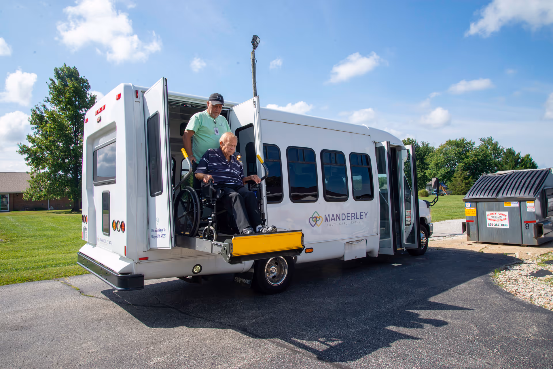 An elderly man in a wheelchair is being assisted by a caregiver as they exit a white shuttle bus equipped with a wheelchair lift. The bus has the logo and name 'Manderley Health Care Center' on its side. The scene is outdoors on a sunny day with green grass and trees in the background.