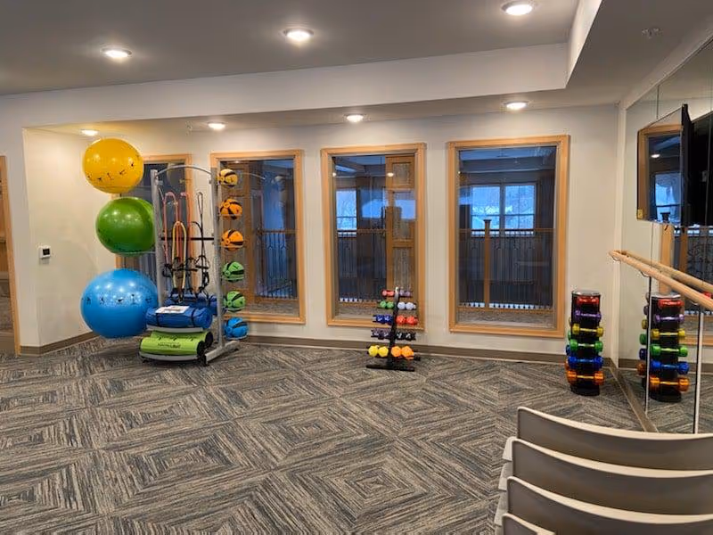 A fitness room with exercise equipment including large colorful exercise balls, various dumbbells on racks, resistance bands, and a mirrored wall with a ballet barre. The room has three large windows with wooden frames and patterned carpet flooring.