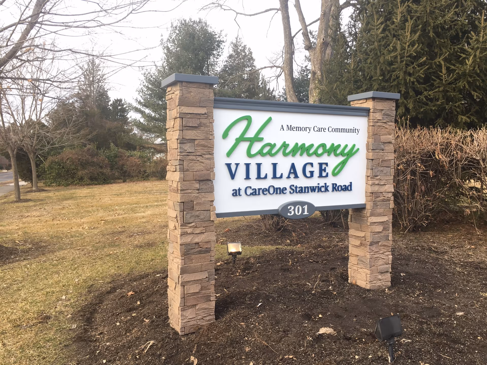 Outdoor stone sign for Harmony Village at CareOne Stanwick Road, a memory care community, with the number 301 displayed below. The sign is surrounded by grass, trees, and bushes in a landscaped area.