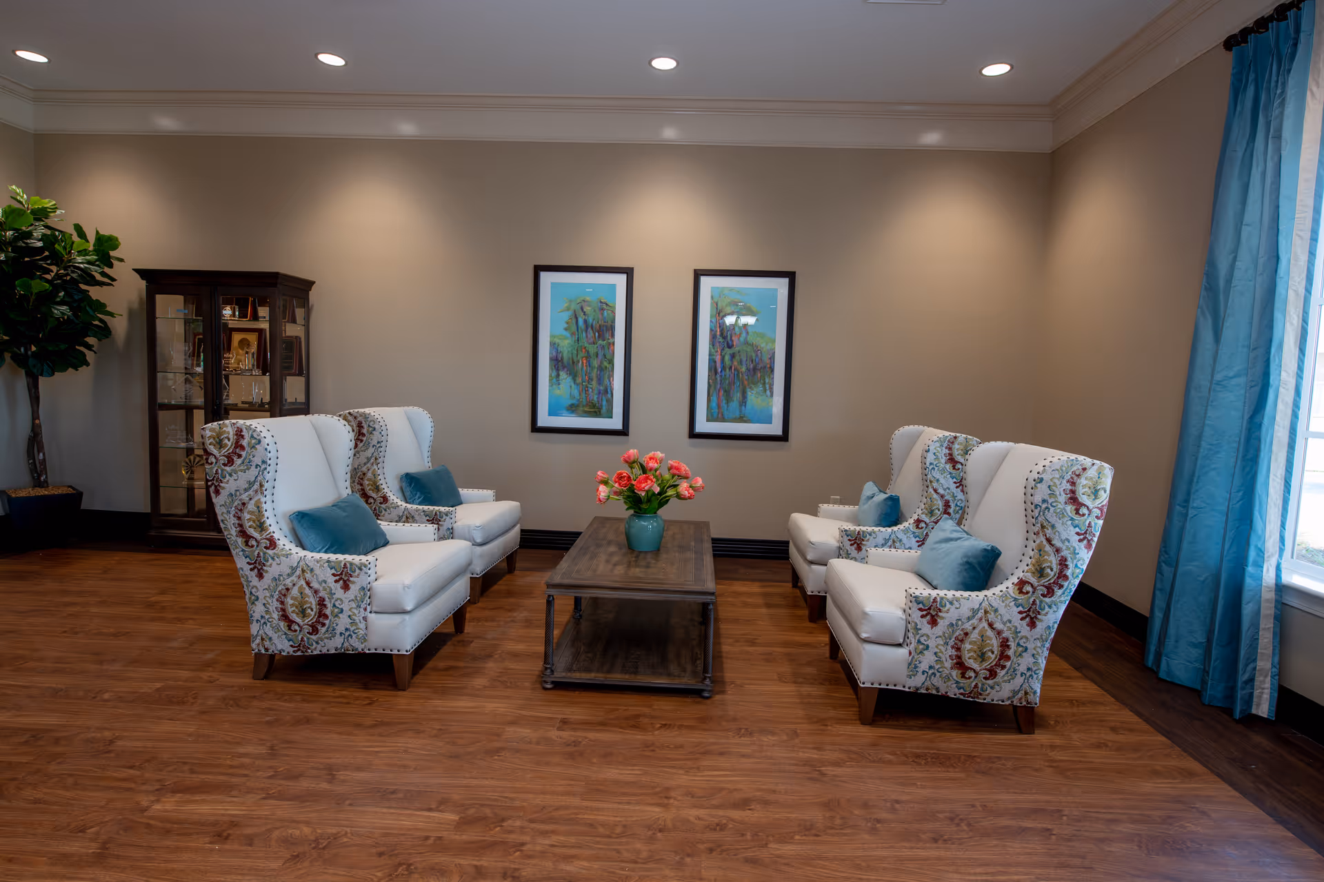 A cozy sitting area with four patterned armchairs arranged around a wooden coffee table with a vase of pink flowers. The room has wooden flooring, beige walls with two framed paintings, a glass display cabinet, a large potted plant, and a window with blue curtains.