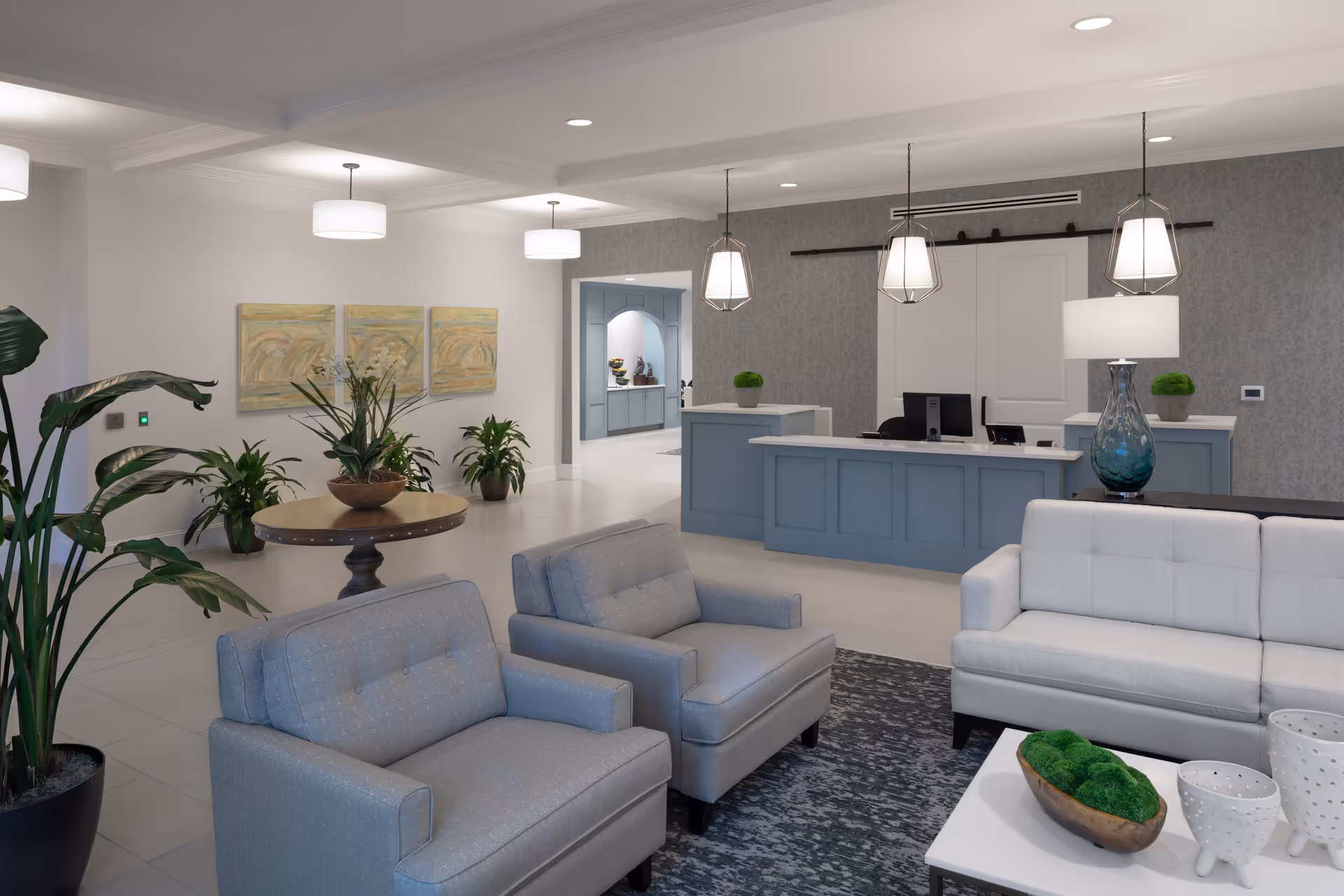 A modern and welcoming senior living facility lobby with comfortable seating including two gray armchairs and a white sofa around a coffee table with decorative items. The reception desk is painted blue with pendant lights hanging above. There are several potted plants and abstract artwork on the walls, creating a calm and inviting atmosphere.