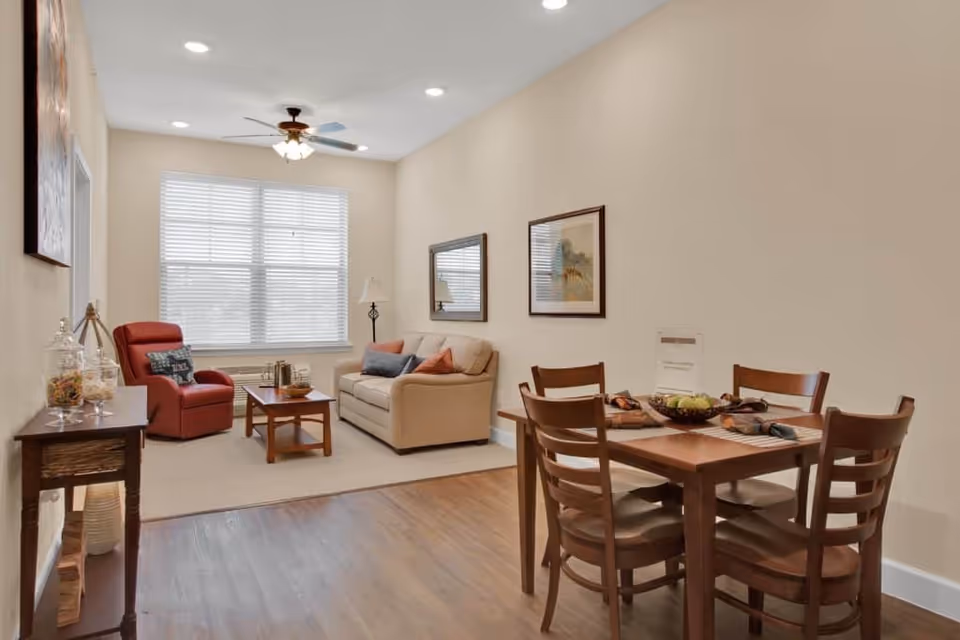Open living and dining area with a beige sofa, red recliner, coffee table, and a wooden dining table with four chairs under recessed lighting.