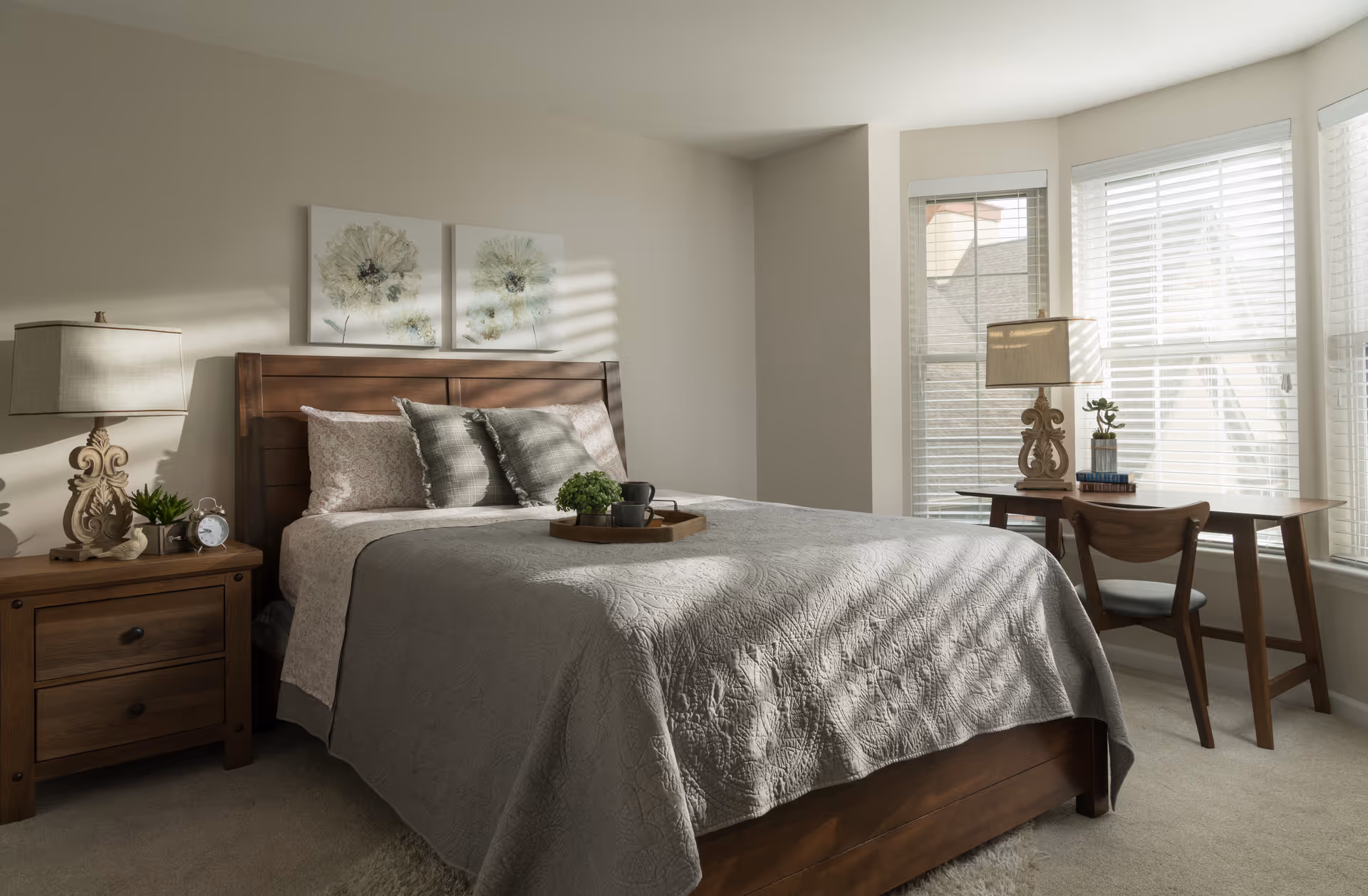 Sunlit bedroom with a wooden bed dressed in gray bedding, matching nightstands and lamps, and a small desk by a bay window.