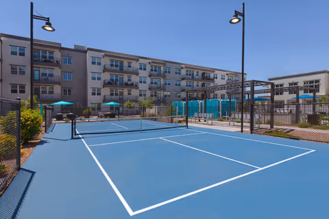 Outdoor tennis court with a net in the middle, surrounded by a chain-link fence. In the background, there is a multi-story residential building with balconies, and several green umbrellas providing shade near seating areas. The sky is clear and blue.