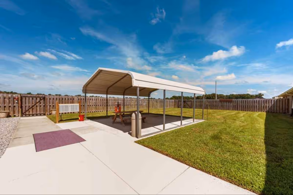 Outdoor covered seating area with a metal canopy, concrete pad, grass lawn, and fence.