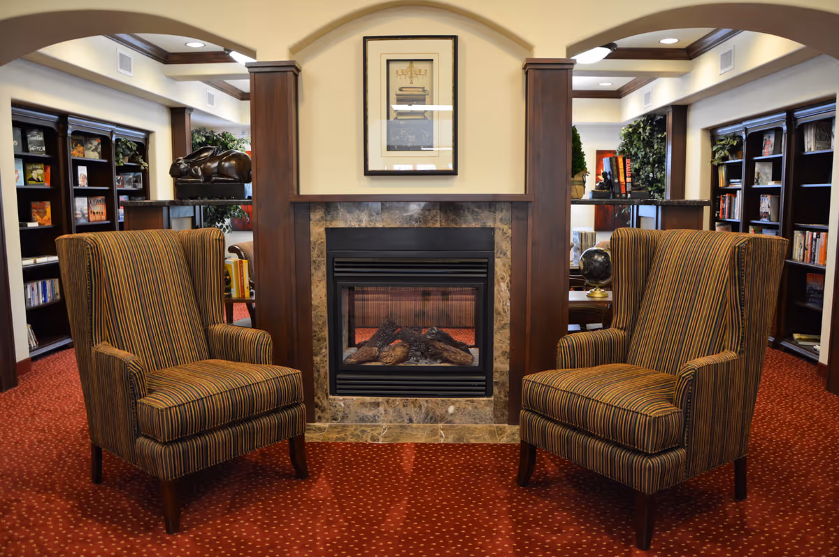 A cozy interior lounge area with two striped armchairs facing a marble-framed fireplace. The room features dark wood accents, built-in bookshelves filled with books, and decorative plants. The carpet is red with a subtle pattern, and a framed picture hangs above the fireplace.