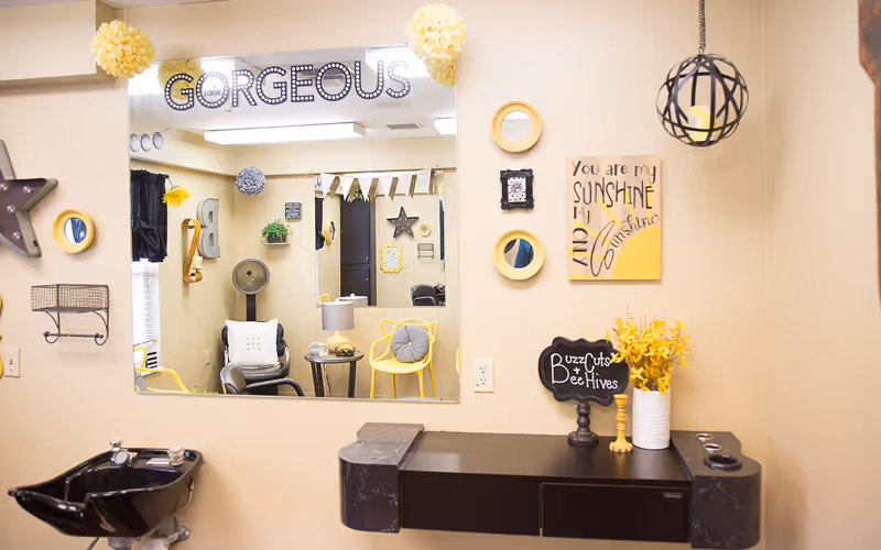 Interior view of a hair salon area with a large mirror reflecting chairs, a fan, and wall decorations. The wall above the mirror has the word 'GORGEOUS' in decorative letters. There are yellow and black decorative items, including a sign that says 'You are my sunshine my only sunshine' and a small chalkboard sign reading 'Buzz Cuts Bee Hives'.