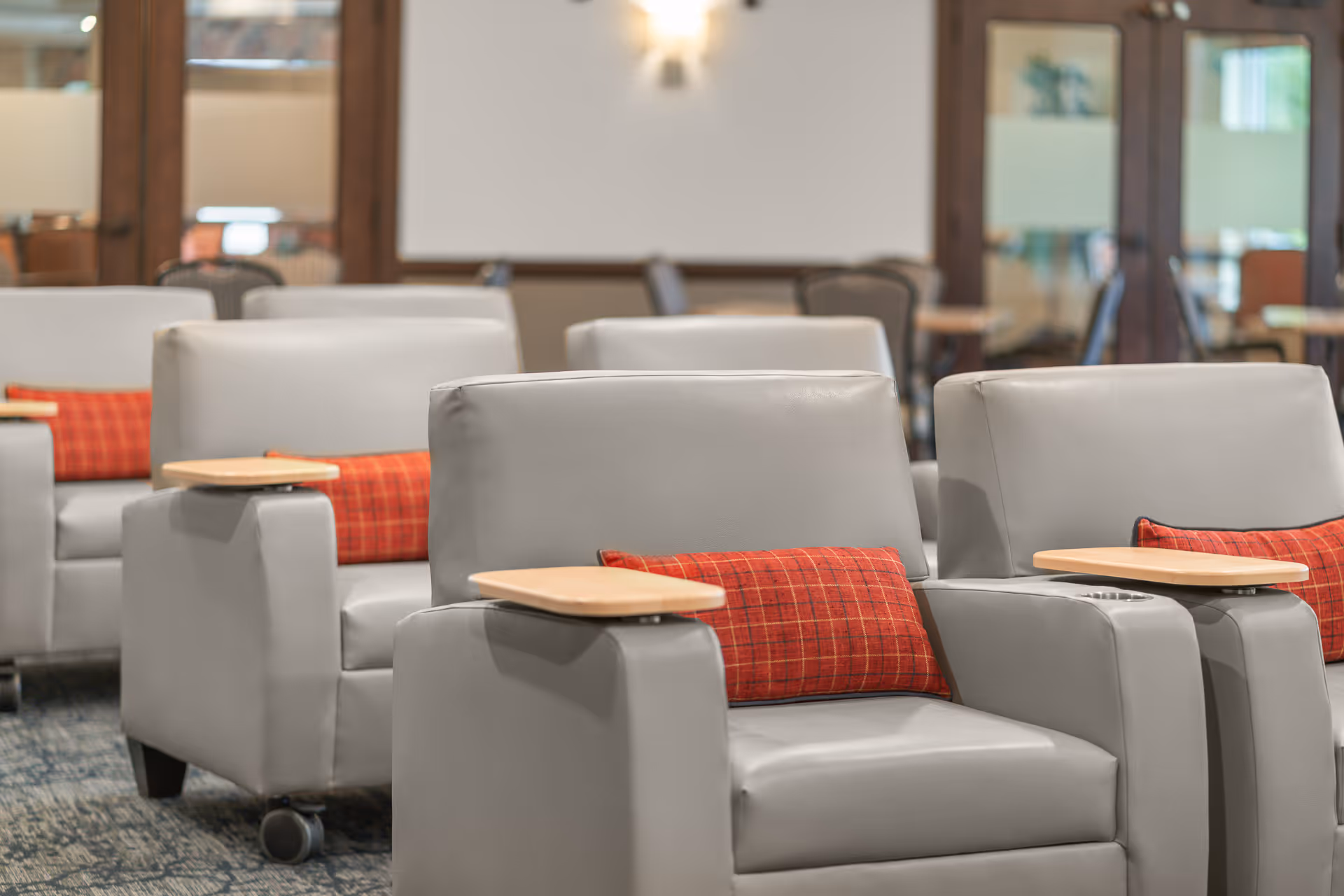 A row of gray leather armchairs with attached wooden trays and red plaid cushions in a well-lit room with carpeted flooring and glass doors in the background.