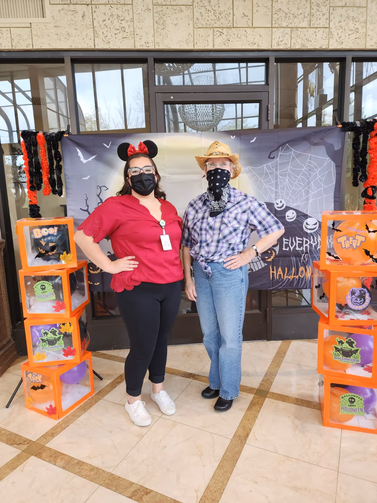 Two women standing indoors in front of a Halloween-themed backdrop. One woman is wearing a red top, black pants, white shoes, a black face mask, and Minnie Mouse ears with a red bow. The other woman is wearing a plaid shirt, blue jeans, black shoes, a black bandana face covering, and a straw cowboy hat. Both have their hands on their hips. There are orange Halloween decoration boxes stacked on either side of them with various Halloween-themed images and balloons inside.
