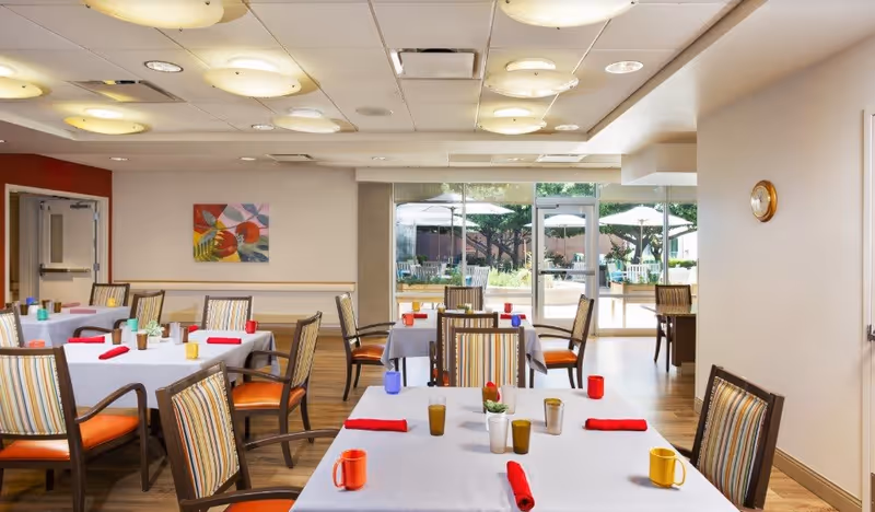A bright dining room with several tables covered in white tablecloths, each set with colorful cups and red napkins. The chairs have striped upholstery and orange seats. Large windows and glass doors at the back open to an outdoor patio area with umbrellas and greenery. The room has a wooden floor, ceiling lights, a wall clock, and a colorful painting on one wall.