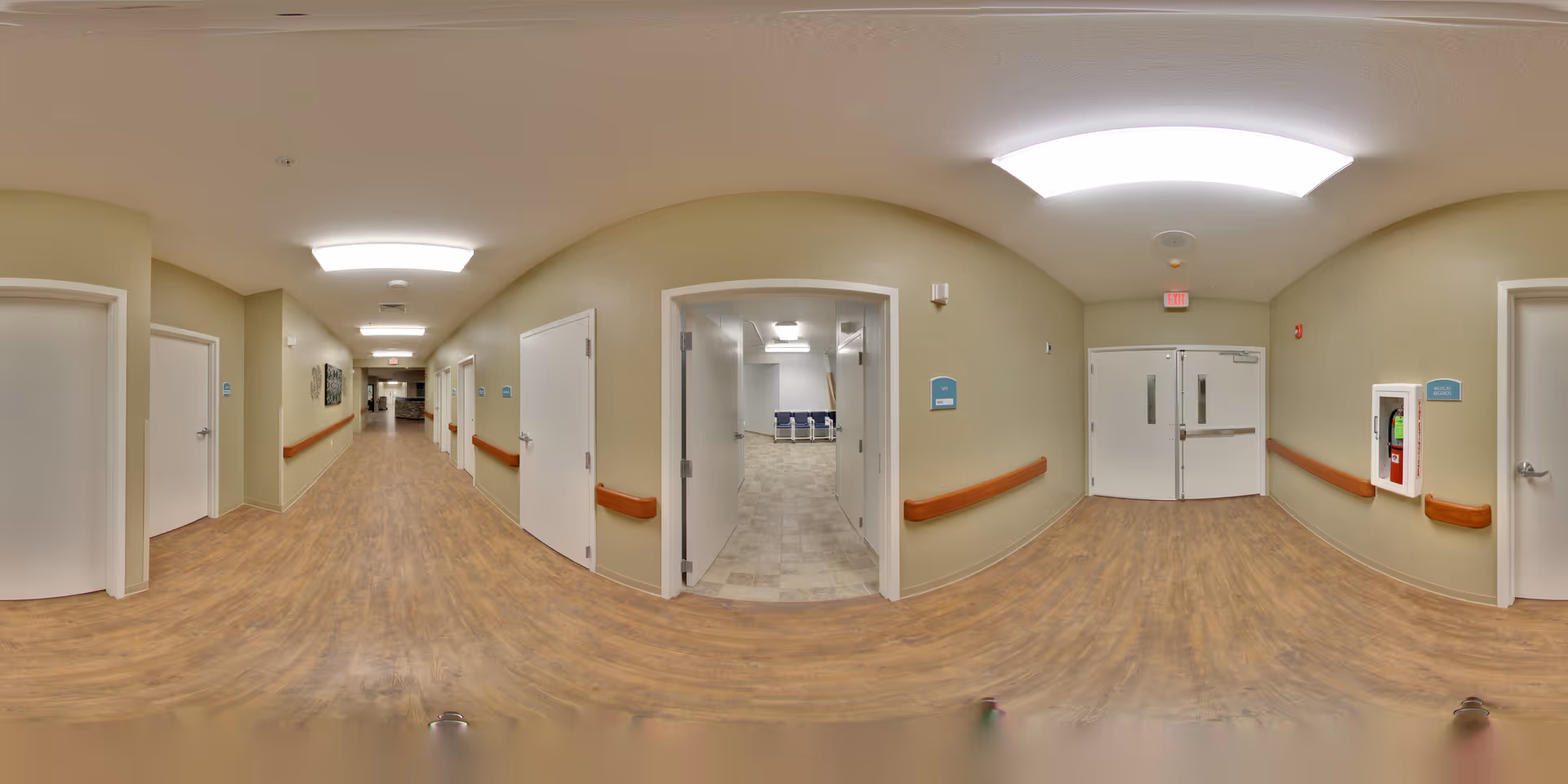 A wide hallway in a senior care facility with light beige walls, wood-look flooring, and white doors on both sides. Handrails run along the walls, and there are ceiling lights providing bright illumination. At the end of the hallway, double exit doors are visible. One open door reveals a room with chairs inside. A fire extinguisher is mounted on the right wall.