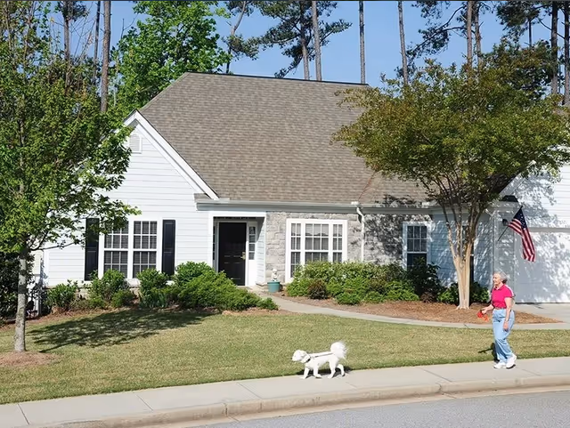 A single-story house with white siding and stone accents, surrounded by green shrubs and trees. A woman in a pink shirt and blue jeans is walking a small white dog on a leash along the sidewalk in front of the house. An American flag is mounted near the garage.