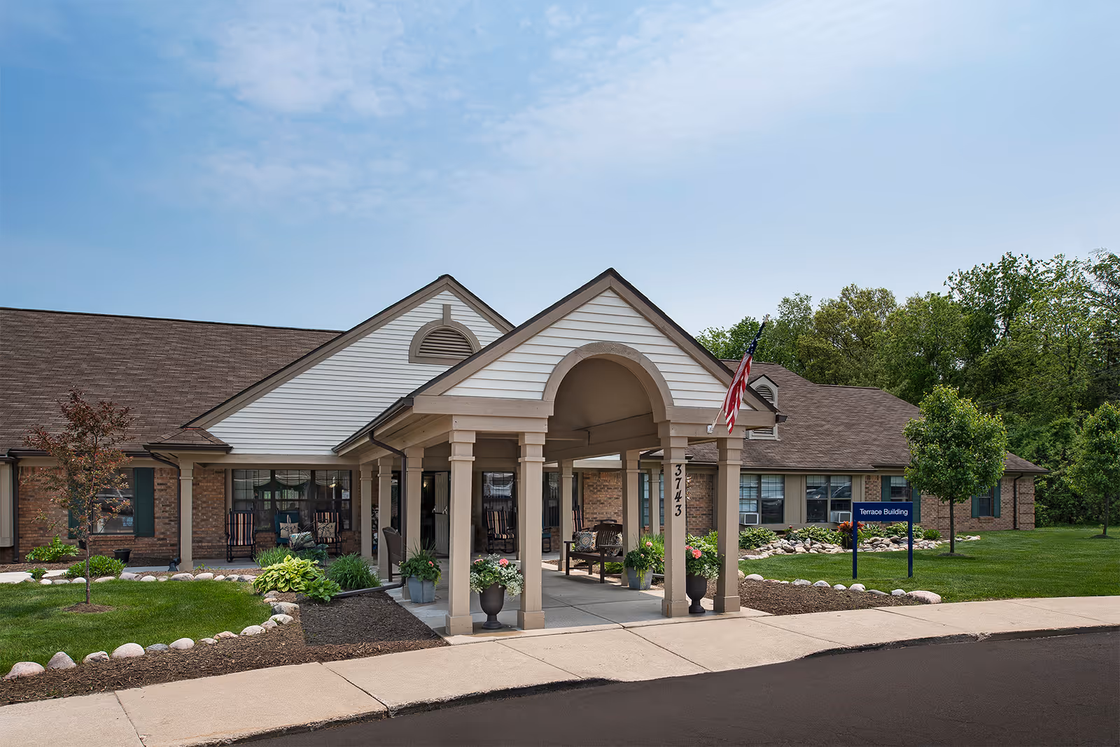Exterior view of a single-story building with a covered entrance supported by columns. The building has a brick and siding facade with a brown shingled roof. There is an American flag hanging from the entrance structure and a sign that reads 'Terrace Building' on the lawn. The surrounding area has green grass, small trees, and landscaping with rocks and plants under a partly cloudy sky.