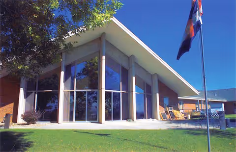 Exterior view of a building with large triangular glass windows and a slanted roof, surrounded by green grass and a flagpole with a flag in front. There are chairs and a small table on the patio area near the building.