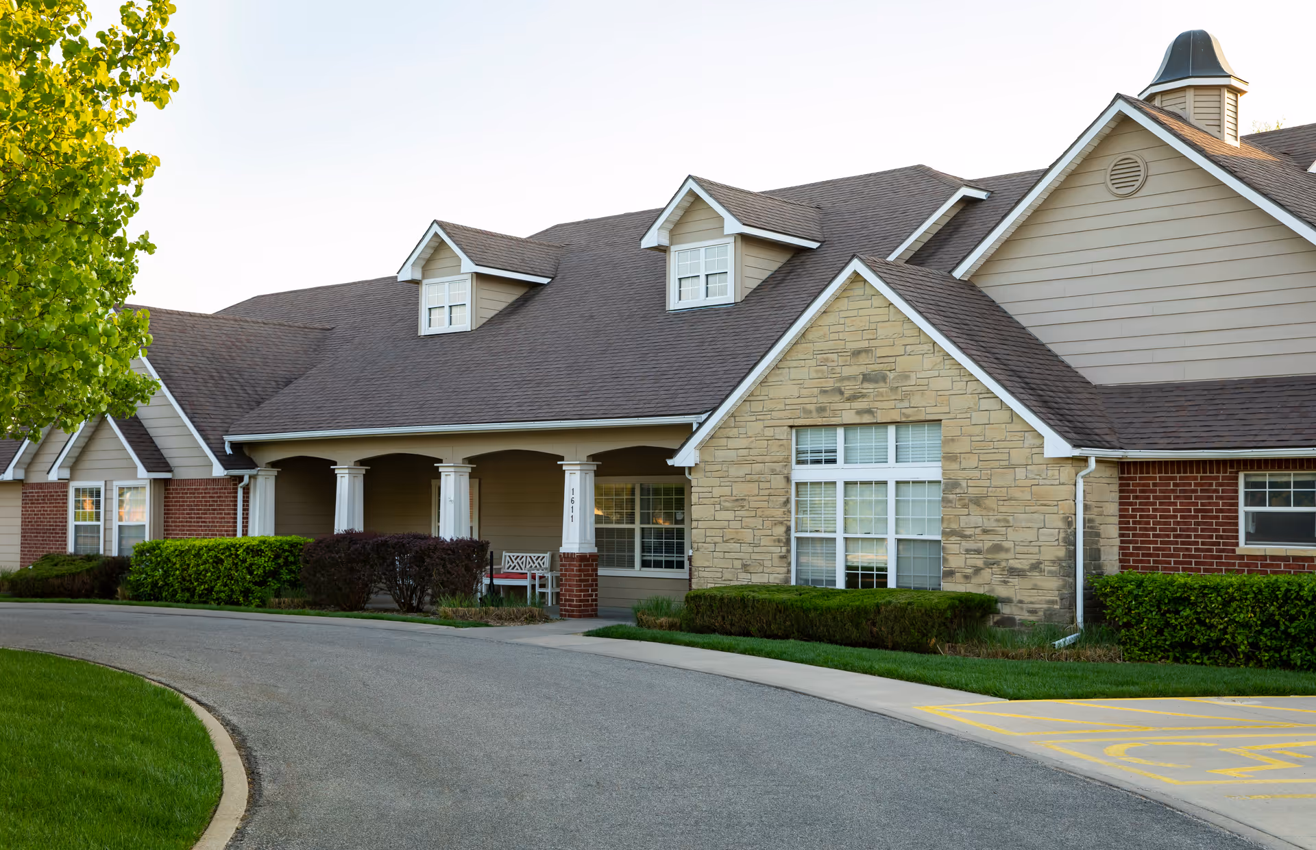 Exterior view of a single-story assisted living facility building with a combination of brick, stone, and siding. The building features a covered porch with white columns, multiple windows, and a curved driveway in front. There is greenery including bushes and a tree near the building.