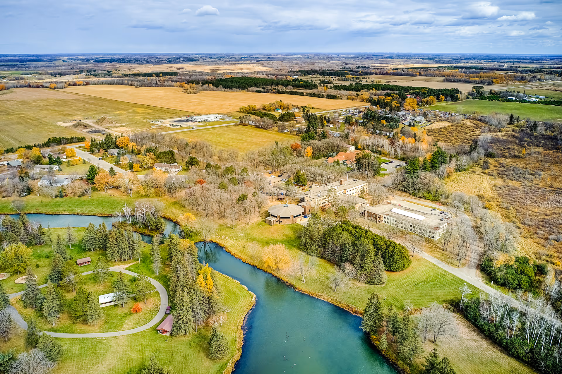 Aerial view of a rural senior living campus with multiple buildings beside ponds, wooded areas, and surrounding farmland.