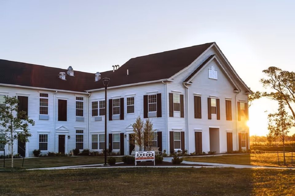Exterior view of a two-story white building with multiple windows and dark shutters, surrounded by a grassy lawn with a few small trees and a white bench in front. The sun is setting in the background, casting a warm glow.