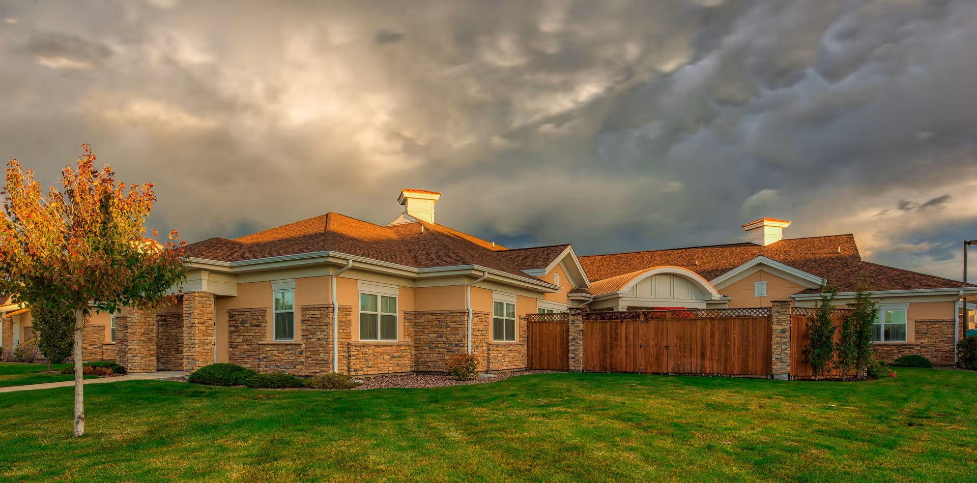 Exterior view of a single-story building with a brown shingle roof, beige walls with stone accents, and several windows. The building is surrounded by a well-maintained green lawn and a small tree with autumn-colored leaves. The sky above is cloudy with a dramatic lighting effect.